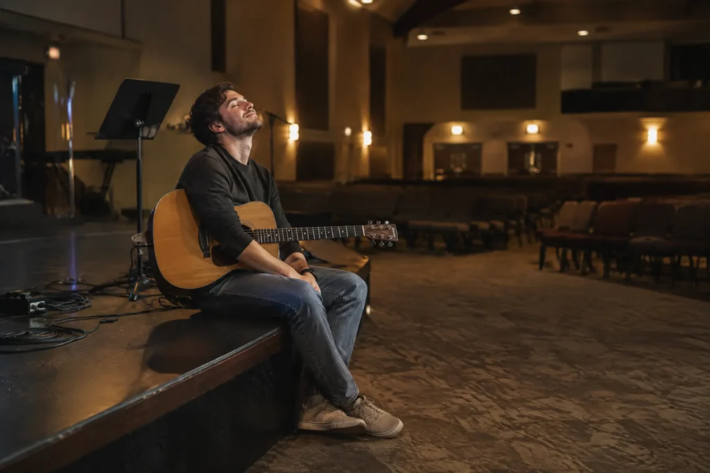 A worship leader in his twenties sitting on the edge of the stage after everyone else has left